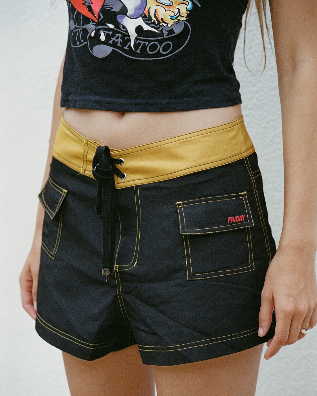 A person wearing black denim shorts with yellow contrast stitching, a yellow waistband, and a black drawstring, paired with a black graphic crop top against a white wall.